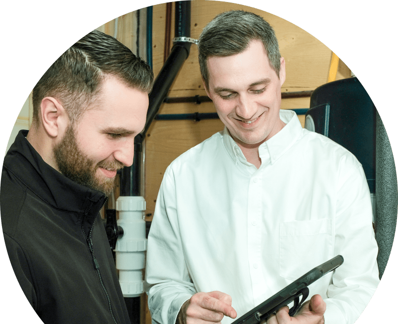 Oosterveld specialist reviewing maintenance plan options with a smiling homeowner using a tablet, inside a utility room with visible piping. Oosterveld specialist reviewing maintenance plan options with a smiling homeowner using a tablet, inside a utility room with visible piping.